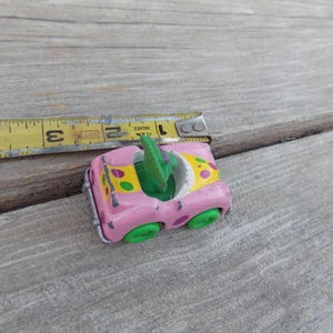 May include: A small, pink toy car with green wheels and a green interior. The car's hood is decorated with yellow and green polka dots. A yellow measuring tape is next to the car, showing inches.