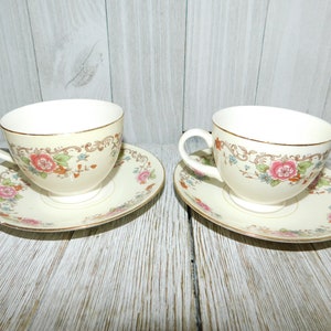 May include: Two cream-colored teacups and saucers with floral designs. The cups and saucers have a gold trim. The floral design includes pink roses, green leaves, and blue accents. The teacups have a curved handle.