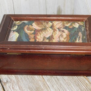 May include: A wooden box with a hinged lid. The lid is decorated with a floral tapestry featuring a variety of colors, including pink, yellow, green, and brown.