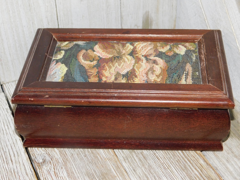 May include: A wooden box with a hinged lid. The lid is decorated with a floral tapestry featuring a variety of colors, including pink, yellow, green, and brown.