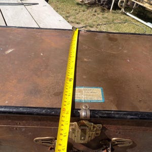 May include: A vintage brown trunk with black trim and brass hardware. A yellow measuring tape shows a length of approximately 19 inches. The trunk has a label that reads "Royal Dutch Airlines".