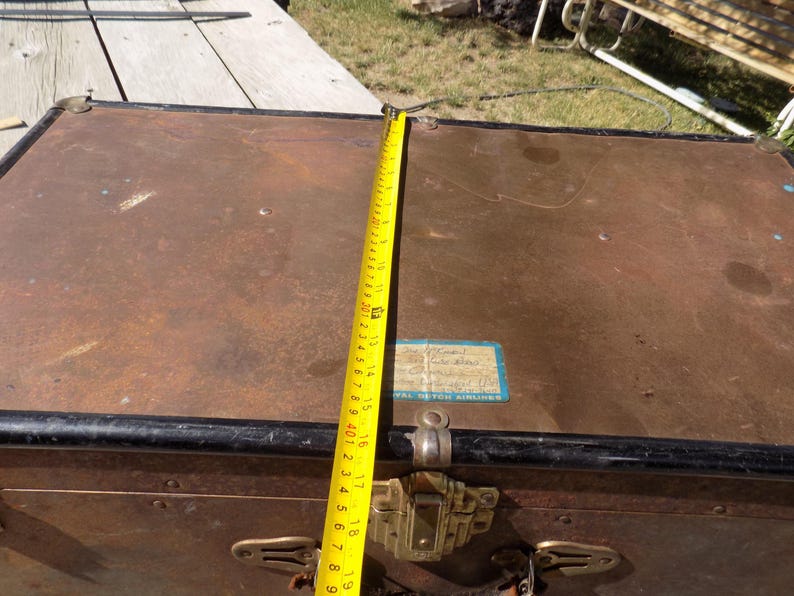 May include: A vintage brown trunk with black trim and brass hardware. A yellow measuring tape shows a length of approximately 19 inches. The trunk has a label that reads "Royal Dutch Airlines".