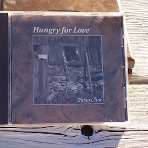 May include: A compact disc in a clear plastic jewel case featuring the title Hungry for Love and the artist name Patsy Cline. The cover art displays a black and white photograph of a rustic wooden building with a stone foundation and a window.