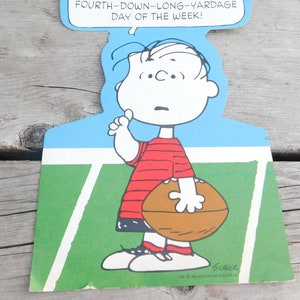 Peut inclure: Une illustration de bande dessin&eacute;e de Charlie Brown tenant un ballon de football sur un terrain vert avec des lignes blanches. Le texte au-dessus de lui dit "Fourth-down-long-yardage day of the week!" et le texte en dessous de lui dit "Schulz".