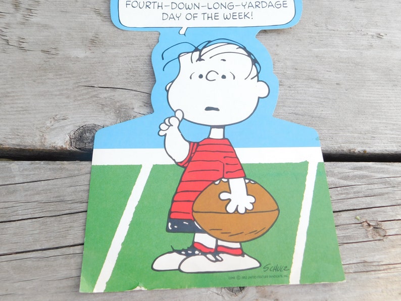 Peut inclure: Une illustration de bande dessin&eacute;e de Charlie Brown tenant un ballon de football sur un terrain vert avec des lignes blanches. Le texte au-dessus de lui dit "Fourth-down-long-yardage day of the week!" et le texte en dessous de lui dit "Schulz".