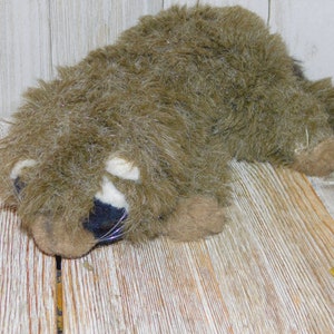 Puede incluir: Un juguete de peluche de mapache con pelaje marr&oacute;n, nariz negra y marcas blancas alrededor de los ojos. El juguete est&aacute; acostado sobre una superficie de madera.