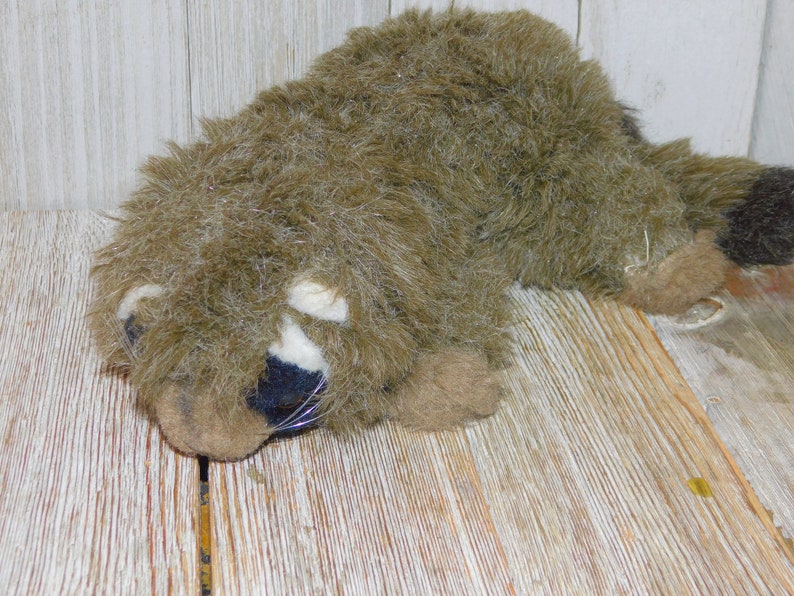 Puede incluir: Un juguete de peluche de mapache con pelaje marr&oacute;n, nariz negra y marcas blancas alrededor de los ojos. El juguete est&aacute; acostado sobre una superficie de madera.