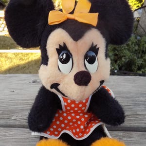May include: A plush Minnie Mouse doll with black fur, a peach-coloured face, and large, expressive eyes. She wears a red and white polka-dot dress and an orange bow. The doll has orange feet and is sitting on a wooden surface.