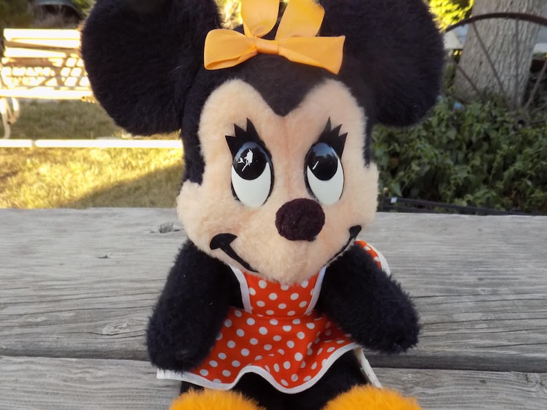 May include: A plush Minnie Mouse doll with black fur, a peach-coloured face, and large, expressive eyes. She wears a red and white polka-dot dress and an orange bow. The doll has orange feet and is sitting on a wooden surface.