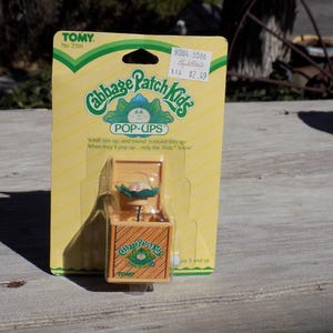 May include: A yellow and green cardboard box with a Cabbage Patch Kids logo and the words "Pop-Ups" printed on it. The box contains a small, yellow toy with a spring mechanism and a Cabbage Patch Kid doll head.