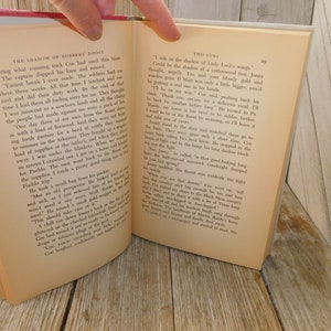 May include: A close-up of an open book with a cream-colored page. The text on the page reads "The Shadow of Borders Roost" and "Two Guns". The text is in black font. The book is open to a page with a paragraph of text.