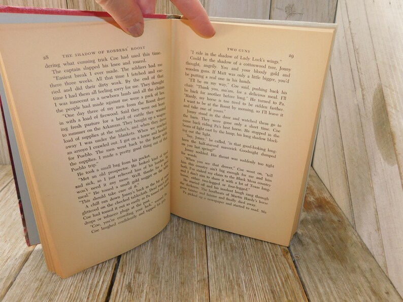May include: A close-up of an open book with a cream-colored page. The text on the page reads "The Shadow of Borders Roost" and "Two Guns". The text is in black font. The book is open to a page with a paragraph of text.