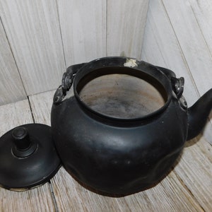 May include: A black cast iron kettle with a lid. The kettle has a rounded body and a spout on the side. The lid has a knob on top.