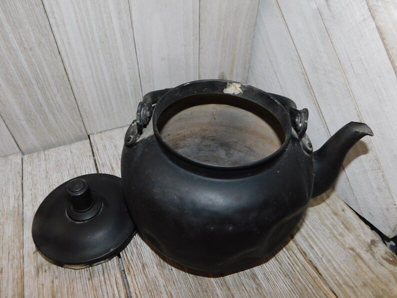 May include: A black cast iron kettle with a lid. The kettle has a rounded body and a spout on the side. The lid has a knob on top.