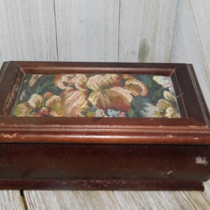 May include: A wooden box with a floral tapestry lid. The box is dark brown and has a rectangular shape. The tapestry features a variety of flowers in shades of pink, yellow, and green.