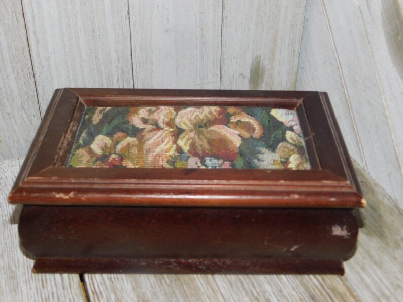May include: A wooden box with a floral tapestry lid. The box is dark brown and has a rectangular shape. The tapestry features a variety of flowers in shades of pink, yellow, and green.
