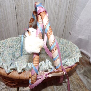Peut inclure: Un panier en osier avec une doublure florale et une poign&eacute;e envelopp&eacute;e de ruban rose et bleu. Un petit animal en peluche blanc est attach&eacute; &agrave; la poign&eacute;e.