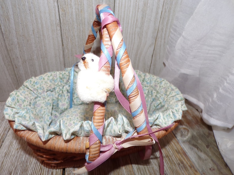Peut inclure: Un panier en osier avec une doublure florale et une poign&eacute;e envelopp&eacute;e de ruban rose et bleu. Un petit animal en peluche blanc est attach&eacute; &agrave; la poign&eacute;e.