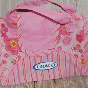 May include: Pink baby changing pad with a floral pattern and vertical stripes. The pad has a handle and a flap with the Graco logo. The floral pattern includes pink, orange, and white flowers.