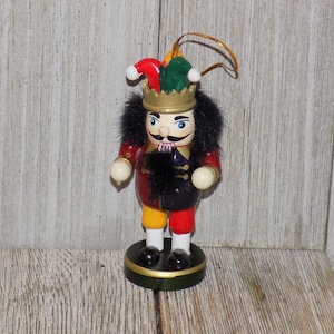 May include: A wooden nutcracker ornament with a red, yellow, and blue costume, a black beard, and a green and red jester hat. The ornament has a gold crown and a gold loop for hanging.