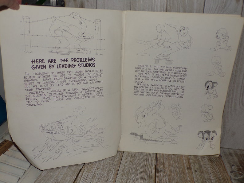 May include: Open book with cartoon sketches and text. The text reads "Here are the problems given by leading studios." The sketches depict various cartoon scenarios, including a man climbing a barbed-wire fence and a man rowing a boat.