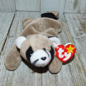 K&ouml;nnte beinhalten: Ein braunes und wei&szlig;es Beanie Baby Waschb&auml;r mit schwarzer Nase und Augen. Das Beanie Baby liegt auf dem Bauch mit eingerolltem Schwanz. Das Beanie Baby hat ein rotes Ty-Etikett mit einem wei&szlig;en Stern und dem Wort "Ty" in wei&szlig;.
