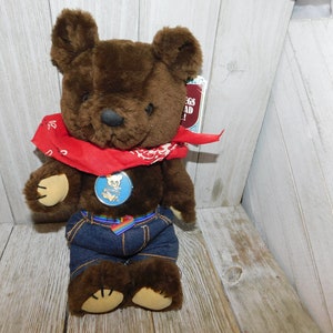 May include: Brown teddy bear wearing a red bandana and blue denim jeans with a rainbow belt. The bear has a round blue badge with a white bear logo on its chest. The bear is sitting on a wooden surface. The text on the tag reads "Hugs Glad!"