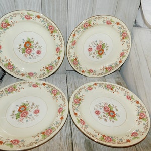 May include: Four cream-colored vintage plates with floral designs. Each plate features a central floral arrangement in orange, pink, and blue, surrounded by a decorative border and gold trim. A classic set for serving.