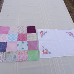 May include: A patchwork quilt with various floral and striped patterns in pink, blue, and purple. Next to it is a white cloth with embroidered floral designs in the corners. The items are placed on a light-colored surface.