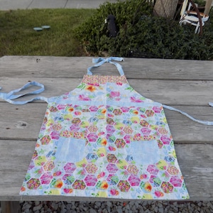 May include: A colorful floral apron with a light blue gingham trim and ties. The apron features a patchwork design with hexagonal patterns in various colors, including pink, yellow, and blue. Two square pockets are visible.