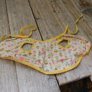 May include: A yellow and white floral print baby bib with yellow trim and two arm holes.