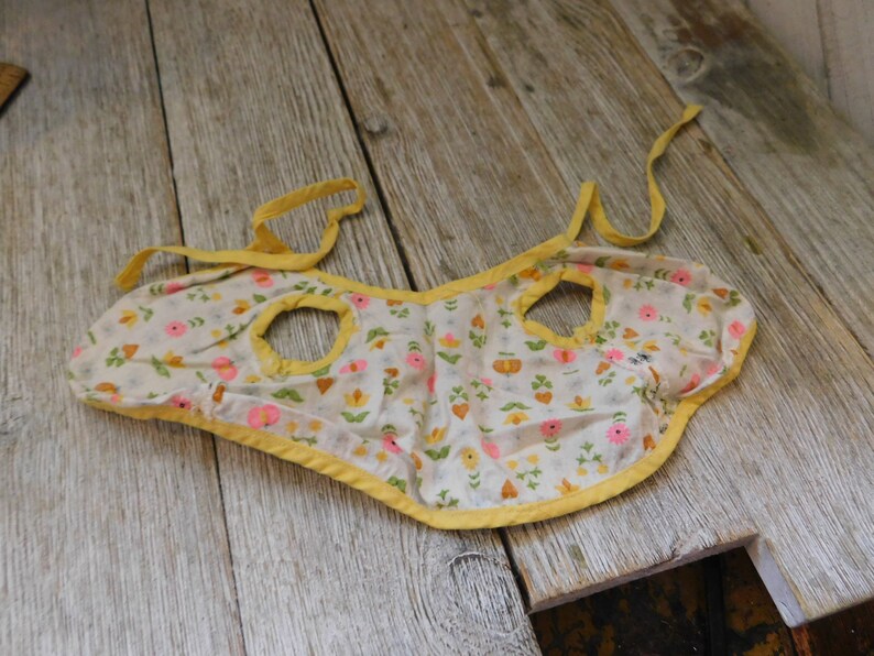 May include: A yellow and white floral print baby bib with yellow trim and two arm holes.