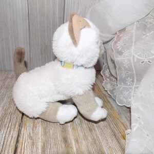 May include: A plush white cat toy with tan ears, paws, and tail. It has a yellow and blue collar. The toy is sitting on a wooden surface, with a white lace curtain in the background.