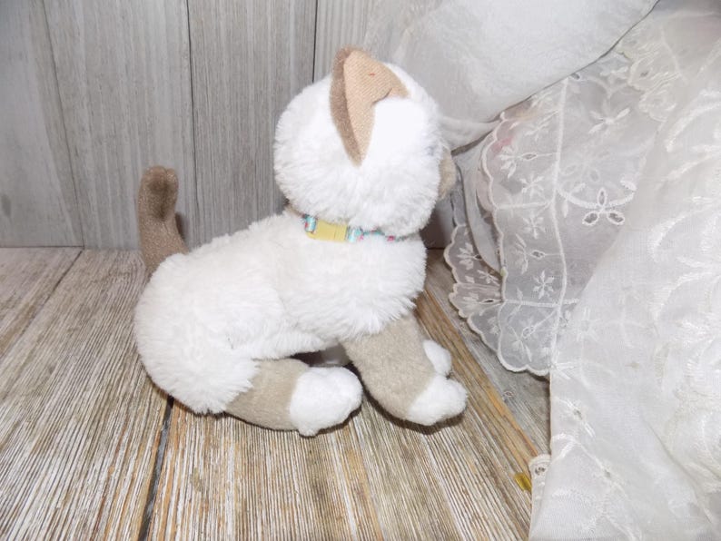May include: A plush white cat toy with tan ears, paws, and tail. It has a yellow and blue collar. The toy is sitting on a wooden surface, with a white lace curtain in the background.