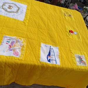 May include: A bright yellow quilt with embroidered patches. The quilt features various designs, including a sailboat, floral wreaths, and figures. The quilt is placed on a weathered wooden surface, showcasing its handmade quality and vibrant color.