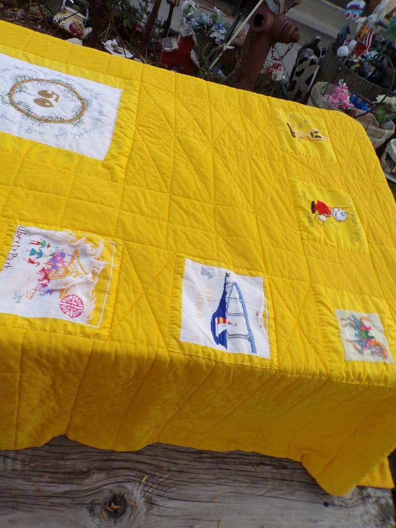 May include: A bright yellow quilt with embroidered patches. The quilt features various designs, including a sailboat, floral wreaths, and figures. The quilt is placed on a weathered wooden surface, showcasing its handmade quality and vibrant color.