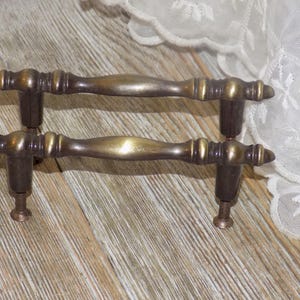May include: Two antique brass drawer pulls. Each pull has a central, curved handle with decorative detailing and dark brown mounting posts. The pulls are mounted with screws. The background is a light wood surface.