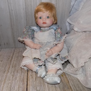 May include: A porcelain doll with blonde hair wearing a blue and white floral dress and white shoes. The doll is sitting on a wooden surface.