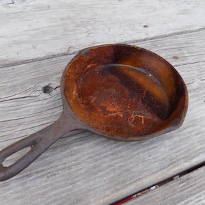May include: An antique cast iron skillet with a single handle and a round cooking surface. The skillet has a dark, rusty brown interior and a gray exterior. The handle has a hole for hanging. The pan is likely vintage.