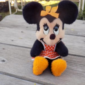 May include: A plush Minnie Mouse doll with black fur, large black ears, and an orange bow. She wears a red and white polka dot dress and has orange feet. The doll is sitting on a weathered wooden surface.