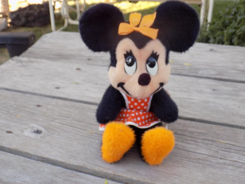 May include: A plush Minnie Mouse doll with black fur, large black ears, and an orange bow. She wears a red and white polka dot dress and has orange feet. The doll is sitting on a weathered wooden surface.