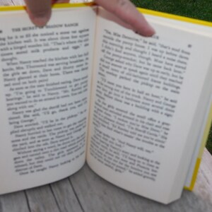 May include: An open book with yellow covers. The pages are filled with text. The book is titled "The Mystery of Shadow Ranch".