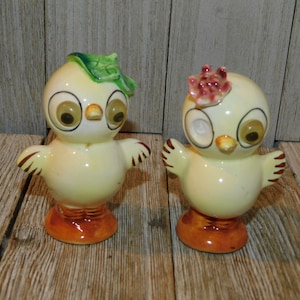 May include: A pair of vintage ceramic bird figurines. One bird has a green leaf on its head, the other has a pink flower. Both have large eyes, outstretched wings, and orange feet. The birds are yellow with brown and orange accents.