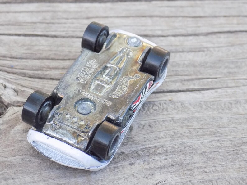 Puede incluir: Un coche de juguete de metal visto desde la parte inferior, con una base met&aacute;lica que muestra el texto CAT-A-PULT, 1998 Mattel y Malaysia. El veh&iacute;culo tiene cuatro ruedas de pl&aacute;stico negro y una carrocer&iacute;a blanca con detalles gr&aacute;ficos en rojo y negro.