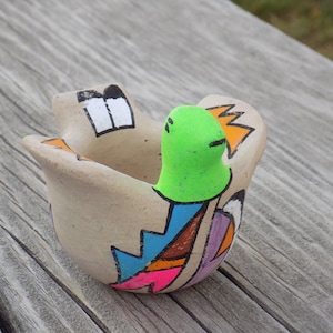 May include: Hand-painted ceramic bird-shaped bowl. The bowl is beige with a green bird head and colorful geometric designs in blue, orange, pink, and purple. The bowl has a unique, artistic design.