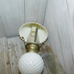 May include: White milk glass oil lamp with a white paper shade. The lamp has a gold metal base and a round, textured globe.