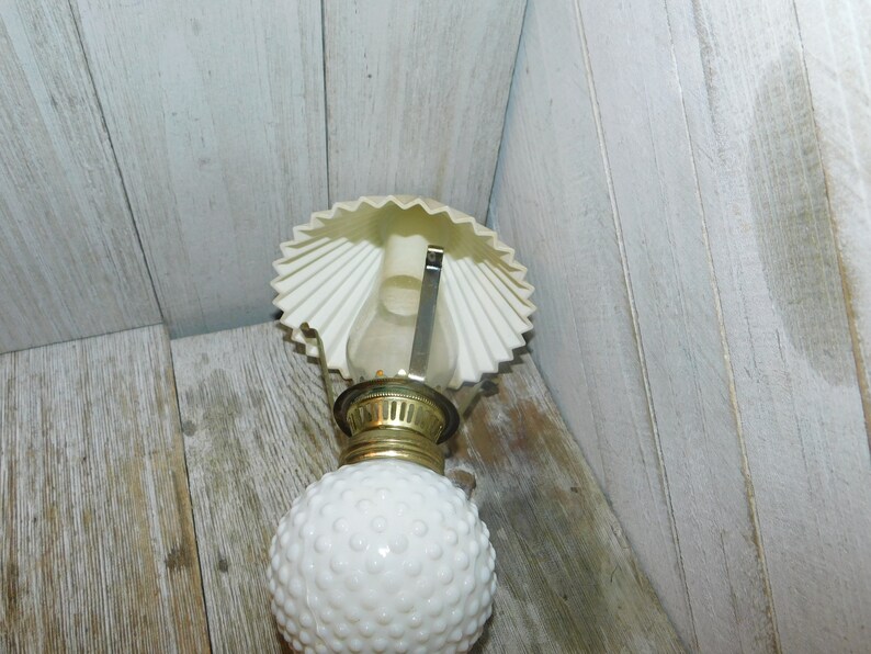 May include: White milk glass oil lamp with a white paper shade. The lamp has a gold metal base and a round, textured globe.