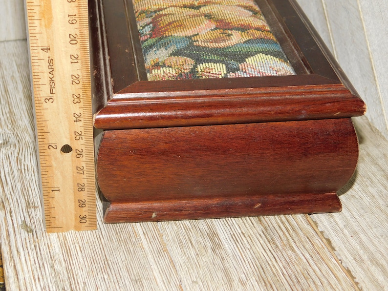 May include: A wooden jewelry box with a hinged lid. The lid is covered in a tapestry fabric with a floral pattern. The box is approximately 9 inches long.