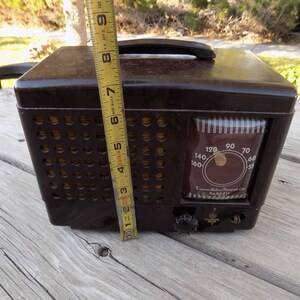 Puede incluir: Una radio Emerson marr&oacute;n vintage con asa, de aproximadamente 20 cm de altura. La radio tiene un dial con n&uacute;meros y una rejilla de altavoz con agujeros circulares. El texto "Emerson Radio/Phonograph Corp. New York USA" es visible.