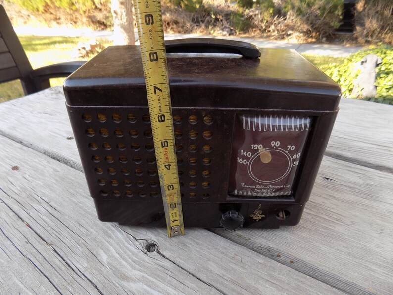 Puede incluir: Una radio Emerson marr&oacute;n vintage con asa, de aproximadamente 20 cm de altura. La radio tiene un dial con n&uacute;meros y una rejilla de altavoz con agujeros circulares. El texto "Emerson Radio/Phonograph Corp. New York USA" es visible.
